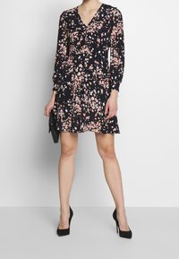 Svart klänning med blommönster i nyanser av rosa och vitt; V-ringning, långa ärmar, utsvängd design, matchas med svarta högklackade skor och clutch.