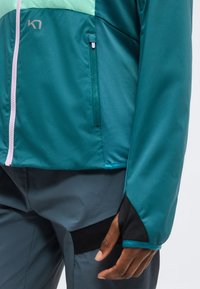 Veste bleu sarcelle et vert clair avec fermeture éclair rose et poche zippée, portée avec un pantalon gris avec des patchs noirs et une main détendue le long du corps.