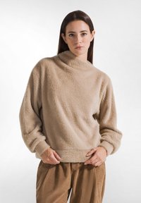 Suéter beige de felpa con cuello alto, tejido texturizado y corte holgado. Mangas largas con puños fruncidos, combinado con pantalones de pana marrones.