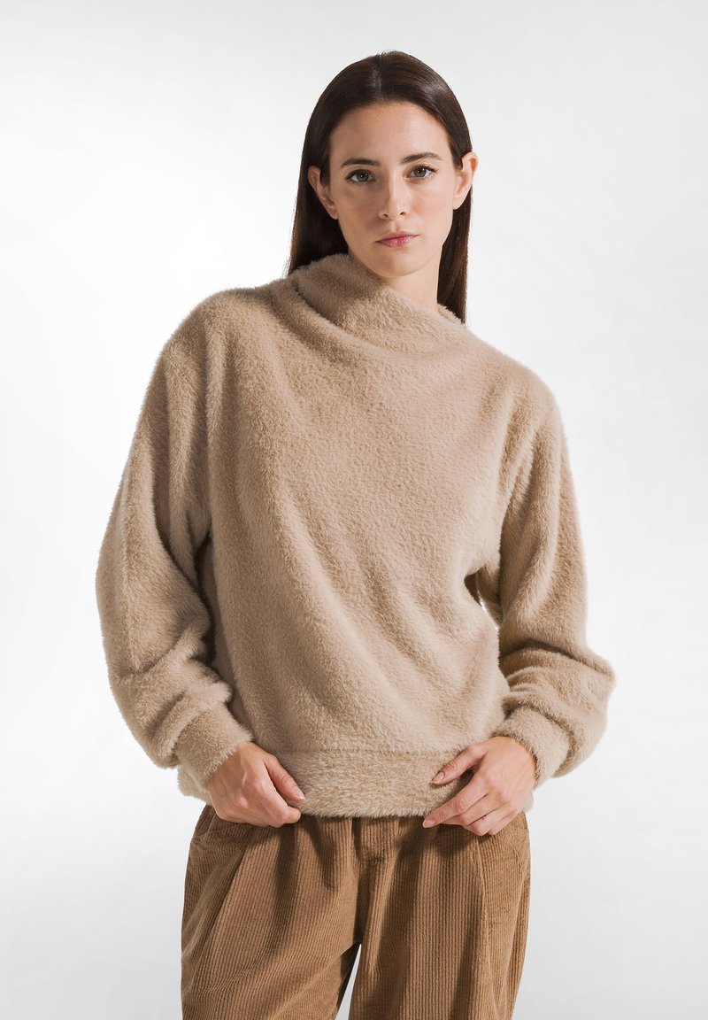 Suéter beige de felpa con cuello alto, tejido texturizado y corte holgado. Mangas largas con puños fruncidos, combinado con pantalones de pana marrones.