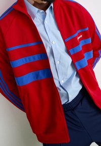 Červená bunda na zip s modrými pruhy, doplněná světle modrou košilí na knoflíky a tmavými šortkami. Látka působí hladce a sportovně.