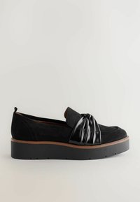 Mustankarvasta loaferit, joissa on kiiltävä musta solki edessä, paksu musta platform-pohja ja ruskeat yksityiskohdat.