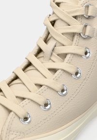 Baskets montantes beiges avec tige en simili cuir, lacets plats, œillets argentés et accents texturés sur la boîte à orteils et le côté.