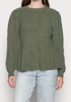 Žena v olivově zeleném svetru s žebrovaným vzorem a nafouklými rameny, v světle modrých džínách, stojící před jednobarevným pozadím.