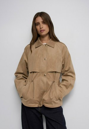 Mujer con cabello largo castaño lleva una chaqueta beige abotonada con cordones y pantalones negros, de pie frente a un fondo gris claro liso.