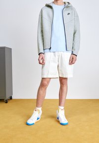 Harmaa vetoketjullinen huppari, vaaleansininen t-paita, valkoiset shortsit ja valkoiset tennarit, joissa sinisiä yksityiskohtia. Sileä pinta, rento urheilullinen muotoilu.
