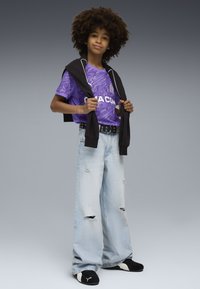 Maglia sportiva a fantasia viola con logo, abbinata a jeans larghi strappati blu chiaro e sneakers nere. Felpa nera poggiata sulle spalle.