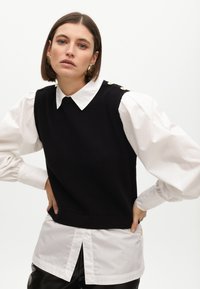 Schwarze Strickweste, die über einem weißen Hemd mit Kragen und Puffärmeln getragen wird, mit goldenen Knöpfen an der Schulter. Glatte, matte Texturen.