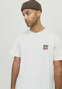Hvid bomulds T-shirt med rund halsudskæring, korte ærmer, prydet med et sort og rødt grafisk logo på venstre bryst. Struktureret stof.