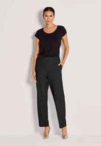 Vrouw staande tegen een witte muur in een zwart t-shirt met korte mouwen, gestructureerde donkergrijze broek, zilveren hakken en hangende oorbellen.