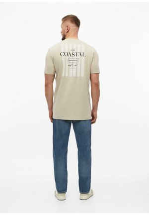 Mężczyzna stoi tyłem, ubrany w beżowy t-shirt w pionowe paski z napisem "Coastal", niebieskie dżinsy i białe trampki.