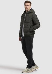 Dunkelgrüne Pufferjacke mit Steppdesign, Kapuze und praktischer Brusttasche mit Reißverschluss. Kombiniert mit dunklen Jeans und hellgrünen Sneakers.