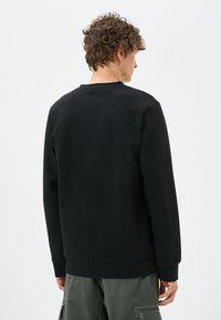 Czarny sweatshirt o fakturowej powierzchni, z okrągłym dekoltem, ściągaczami na rękawach i w pasie. Noszony z luźnymi zielonymi spodniami cargo.