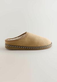 Beige suede slipper with a rounded toe, soft lining, and a braided black-and-white trim around the sole. Closed back, slip-on design.