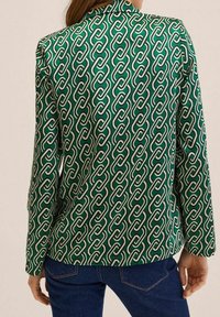 Grüner Blazer mit Muster in Form einer Kettenverbindung in Weiß. Der Stoff erscheint glatt, hat eine taillierte Passform, lange Ärmel und einen Kragen.
