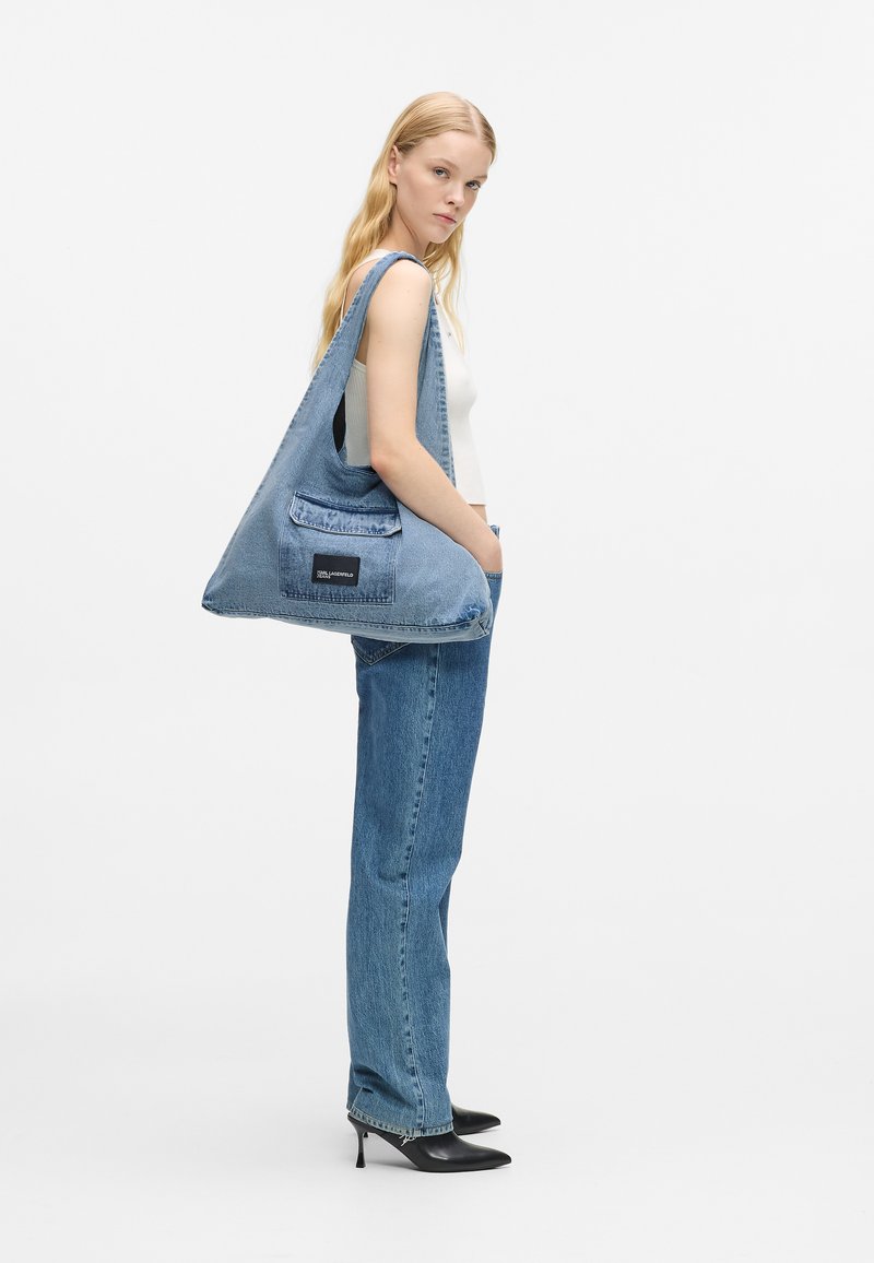 Mulher vestindo uma blusa branca de alças, jeans azuis, sapatos de salto alto pretos, carregando uma grande mala de ombro de denim azul com um bolso frontal decorado.