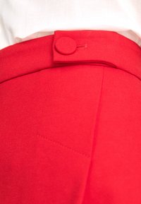 Červené nohavice majú hladkú textúru a zapínanie na gombík v páse. Obsahujú jemný vyšitý detail vrecka na boku.