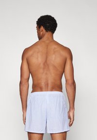 Stripete svømmeshorts i lyseblått og hvitt, laget av lett materiale. Har elastisk midje og en avslappet passform. Bakutsikt vist.