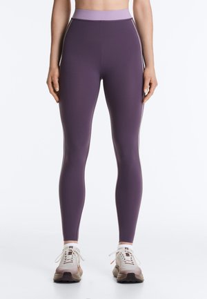 Woman wearing high-waisted purple leggings with light purple waistband and white athletic sneakers standing against plain background.