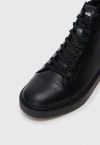 Sorte høyhælte sneakers laget av glatt skinn, med rund tåhette, svarte snørebånd og en logo på tungen.