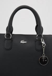 Sac à main noir texturé avec poignées arrondies, logo crocodile en argent, et un porte-clés rond décoré d'un motif de polo, attaché à la poignée.