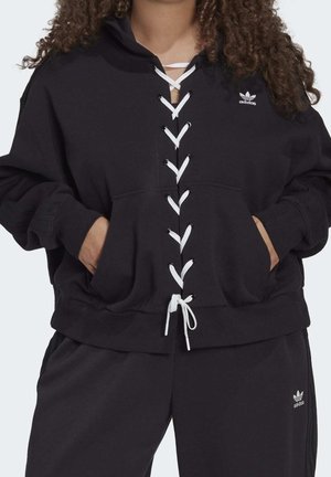 Femme portant un sweat à capuche Adidas noir avec fermeture à lacets blanche à l'avant, poche kangourou, et pantalon Adidas noir assorti.