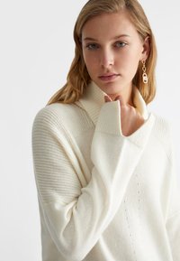 Maglione color crema con collo alto, presenta un motivo a maglia testurizzato e dettagli a coste. Il tessuto morbido si drappeggia attorno alle braccia di chi lo indossa.