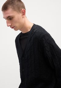 Mustakangasneule, jossa on teksturoitu timanttikuvio, V-pääntie, viisi nappia ja pitkät hihat. Puettu mustan paidan päälle.