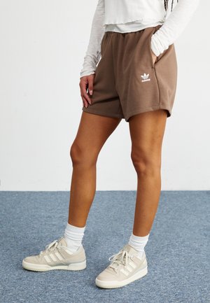 Personne portant un short Adidas marron, un haut à manches longues blanc, des chaussettes blanches montantes et des baskets Adidas beiges, se tenant sur un tapis bleu.