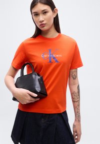 Πορτοκαλί βαμβακερό μπλουζάκι με μπλε λογότυπο "CK" και κείμενο "Calvin Klein", συνδυασμένο με μια μικρή μαύρη τσάντα. Το μοντέλο φοράει τζιν φούστα.
