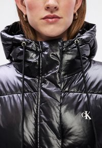 Melnā pūkainā jaka ar spīdīgu apdari, augstu apkakli un aukliņu. Iekļauts neliels sudraba logotips uz krūtīm. Kvadrātveida tekstūra.