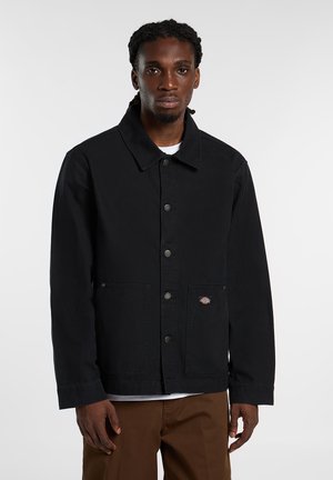 Schwarze Denimjacke mit spitzem Kragen, Knopfleiste vorne, zwei Seitentaschen und einem Logopatch auf der linken Tasche.