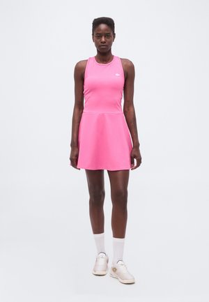 Žena stoji okrenuta naprijed noseći ružičastu tenisku haljinu bez rukava, bijele čarape i svijetle tenisice na jednobojnoj pozadini.