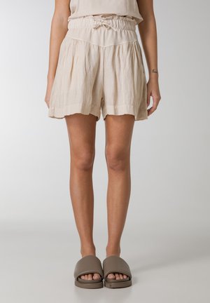 Beige, linen shorts with an elastic waistband and tie detail, featuring a flared shape and ruffled hem. Paired with brown slide sandals.