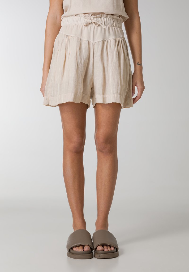 Pantaloni corti beige in lino con cintura elasticizzata e dettaglio a fiocco, caratterizzati da una forma svasata e orlo arricciato. Abbinati a sandali a scivolo marroni.