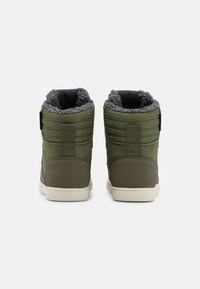 Par de zapatillas altas verdes con suelas blancas y forro de lana gris, vistas desde atrás sobre un fondo liso.