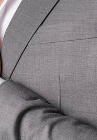 Veste de costume grise avec une texture subtile. Présente une poche unique et une coupe ajustée. Gros plan sur le tissu et le détail de la manche.