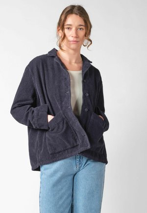 Frau trägt eine dunkelblaue Cordjacke mit Knöpfen und Taschen sowie hellblaue Jeans, steht vor einem einfarbigen hellen Hintergrund.