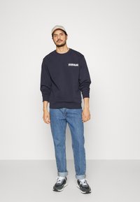 Felpa blu navy con polsini e bordo a coste, motivo grafico frontale. Abbinata a jeans azzurri e sneakers nere. Cappellino indossato al contrario.