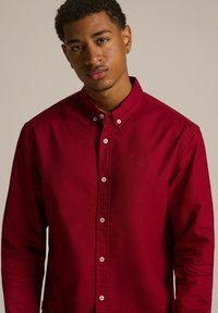 Czerwona koszula z długim rękawem, wykonana z bawełny, z białymi guzikami i haftowanym logo na piersiach. Gładka faktura.