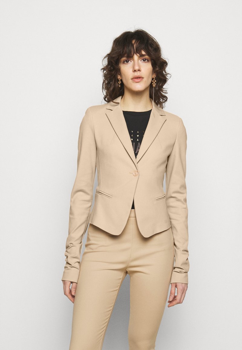 Un blazer beige ajusté avec une fermeture à un seul bouton, des revers crantés et deux poches avant, porté sur un haut noir et un pantalon assorti.