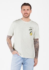Man met tatoeages op beide armen, draagt een lichtbeige t-shirt met een citroen tak grafiek en lichtblauwe jeans, hand in de zak.