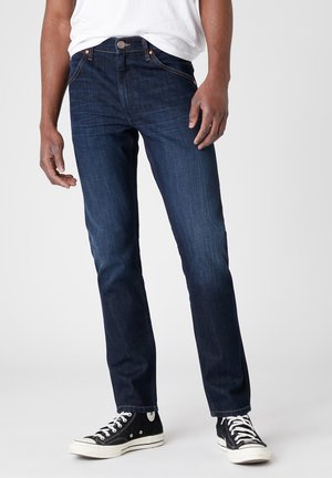 Mann, der dunkelblaue, schlanke Jeans, ein weißes T-Shirt und schwarze High-Top-Sneaker trägt, steht vor einem einfarbigen Hintergrund.