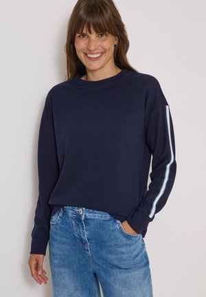 Vrouw die lacht, draagt een donkerblauw shirt met lange mouwen met witte strepen op de mouwen en een blauwe spijkerbroek, staand met één hand in de zak.