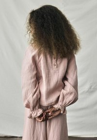 Blouse en coton rose pâle avec une surface texturée, des manches longues et bouffantes, et un bord en dentelle aux poignets, associée à un vêtement coordonné.