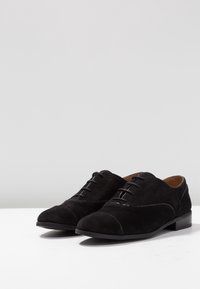Scarpe in suede nere con lacci, punta tonda, dettagli lucidi, tacco piatto e cuciture decorative sulla punta e sui lati.