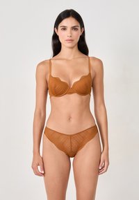 Mujer con cabello largo y oscuro vestida con un conjunto a juego de sujetador y ropa interior de encaje de color óxido, de pie contra un fondo claro y liso.