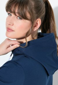 Felpa con cappuccio blu navy dal tessuto morbido. Il modello tocca il colletto, mettendo in evidenza i dettagli delle cuciture e i lacci.