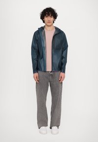 Translucent navy regnjakke med hætte, brystlomme og grå detaljer, båret over en lyserød skjorte og grå jeans. Hvide sneakers.