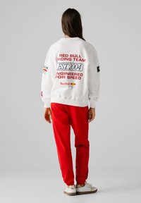 Γυναίκα που φοράει λευκό φούτερ Red Bull Racing με την επιγραφή «EST. 2004» και κόκκινο παντελόνι, στέκεται με την πλάτη προς τα εμπρός μπροστά σε απλό γκρι φόντο.